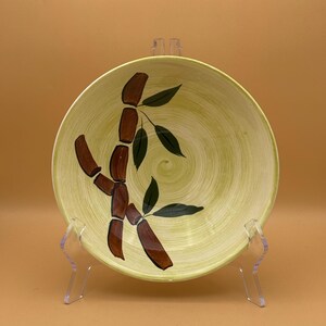 May include: A round, vintage ceramic plate with a light green background and a brown bamboo design with green leaves. The plate is displayed on a clear plastic stand against a light orange background.