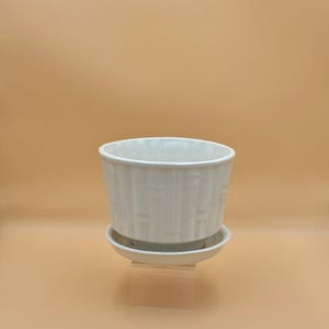 May include: A white ceramic planter with a subtle vertical bamboo-like texture on the exterior. The cylindrical pot features a wide, open top and sits securely on a matching circular saucer base, creating a cohesive and functional plant container.