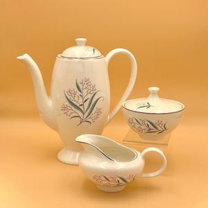 May include: A white ceramic tea set consisting of a tall teapot with a lid, a lidded sugar bowl, and a creamer. Each piece features a delicate botanical pattern of thin green leaves and small pink flowers, accented with a thin silver rim.