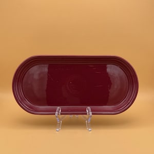 May include: A burgundy-colored, oval-shaped serving platter. The platter has a glossy finish and a raised rim. It is displayed on a clear stand against a solid orange background. The platter is approximately 12 inches long.