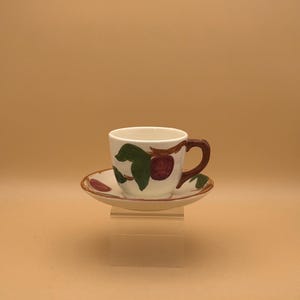 May include: A white ceramic teacup and saucer set. The cup has a brown handle and is decorated with green leaves and red fruit. The saucer has a matching design. The set is displayed on a clear acrylic stand against a tan background.
