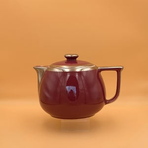 May include: A deep burgundy ceramic teapot featuring a glossy finish, a matching lid with a small knob, and metallic gold-toned accents along the rim, spout, and handle. The teapot has a rounded body and a structured, angular handle design.