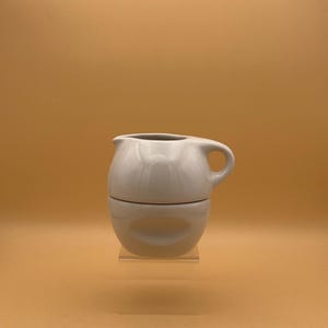 May include: A white ceramic creamer and sugar bowl set. The creamer has a handle and spout, and the sugar bowl has a matching rounded shape. The set is displayed on a clear acrylic stand against a solid orange background.