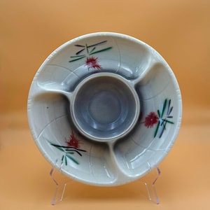 May include: A round ceramic serving dish with a central circular bowl surrounded by three separate compartments. The off-white surface features a subtle crackle glaze and is decorated with three painted motifs of red flowers and green leaves.