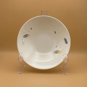 May include: A white ceramic bowl with a wide rim, decorated with small, abstract shapes in black, gray, and yellow. The bowl is displayed on a clear plastic stand against a light brown background.