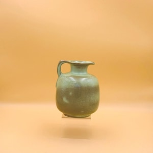 May include: A small ceramic pitcher with a rounded body and a single handle. The vessel features a matte finish with a gradient of muted green and earthy brown tones, transitioning from a lighter green at the top to a darker brown at the base.