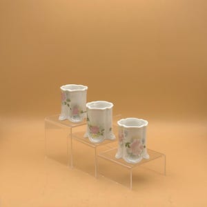 May include: Three white porcelain bud vases with scalloped rims and floral designs. The vases are arranged on clear acrylic risers, creating a tiered display. Each vase features pink and green floral patterns.