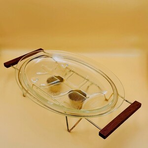 May include: An oval clear glass serving dish with a textured bottom surface, held within a gold-toned metal wire frame stand featuring two rectangular dark brown wooden handles on each side for easy carrying and serving of food items.