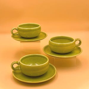 May include: Three matching olive green ceramic tea cups and saucers. Each cup features a rounded bowl shape with a single side handle, paired with a circular saucer. The set displays a smooth, glossy finish throughout the entire surface area.