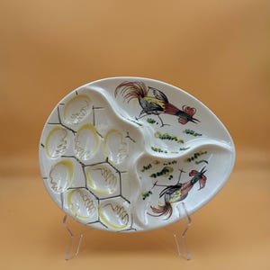 May include: An oval ceramic serving platter with a divided design. One side features six circular indentations outlined in yellow with black sketch-like details. The other side displays two hand-painted roosters in red, black, and yellow tones.