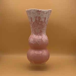 May include: A pink ceramic vase with a unique hourglass shape. The vase has a bulbous base and a wider top, with a textured white and pink pattern on the upper portion. The vase is displayed on a clear stand.