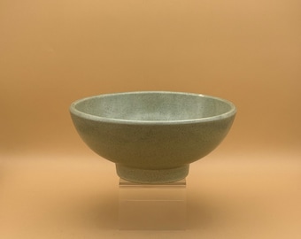 May include: A light green ceramic bowl with a speckled texture. The bowl has a rounded shape and sits on a small, pedestal base. The bowl is displayed against a light brown background.