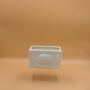 May include: A rectangular off-white ceramic planter with a smooth, glossy finish. The front face features a centered, raised oval design element. The vessel has an open top and straight, vertical sides, providing a clean, functional structure.