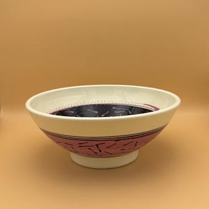 May include: A white ceramic bowl with a pink and black geometric design. The bowl has a wide rim and a rounded base.