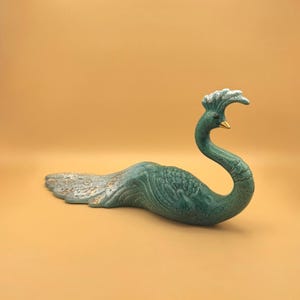 May include: A ceramic peacock figurine in shades of green, with a textured body and a curved neck. The peacock has a gold-colored beak and a detailed tail. The figurine is a decorative item.
