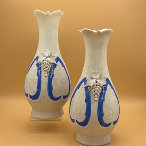May include: Two white ceramic vases with blue accents and grape cluster designs. The vases have a tall, slender shape with a flared top. The grape clusters are white, and the leaves are embossed. The vases are displayed on a neutral background.