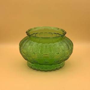 May include: A green glass vase with a textured, patterned design. The vase has a rounded body and a scalloped rim.