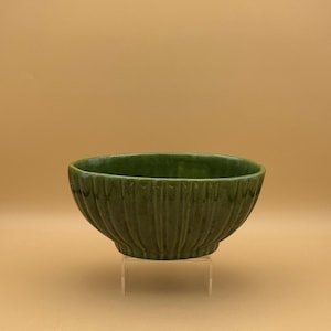 May include: A green ceramic bowl with a ribbed exterior. The bowl is oval-shaped and sits on a clear acrylic stand. The bowl's color is a deep, forest green, and it has a glossy finish.