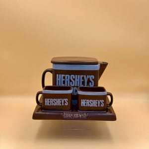 May include: A brown ceramic Hershey's tea set featuring a teapot with a lid, two square mugs, and a rectangular serving tray. Each piece is brown with light blue trim and displays the white Hershey's logo. The tray also features the logo.