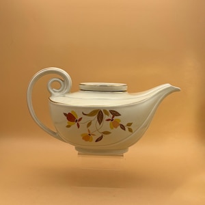 May include: Cream-colored ceramic teapot with a curved handle and spout. The teapot features a floral design in shades of red, yellow, and brown. The rim and handle are accented with gold trim. A decorative piece for serving tea.