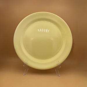 May include: A pale yellow ceramic plate with a wide rim. The plate is displayed on a clear plastic stand against a solid tan background. The plate's surface has a glossy finish.