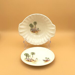 May include: Two-piece vintage ceramic dish set. The set includes a scalloped-edge bowl and a round plate. Both pieces feature a desert scene with palm trees and camels in shades of green, brown, and tan against a white background.
