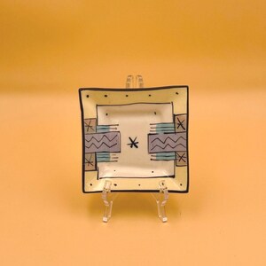 May include: A square ceramic dish with a cream-colored base, featuring a black rim and black polka dots. The center displays a black asterisk symbol flanked by two rectangular blocks decorated with horizontal purple and teal wavy lines.