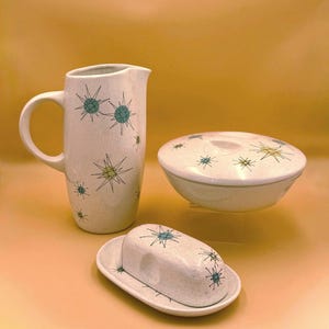 May include: A ceramic kitchen set featuring a tall pitcher, a lidded bowl, and a butter dish. Each piece is off-white with a speckled finish and decorated with teal and yellow starburst patterns consisting of thin lines and small solid circles.