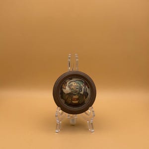 May include: A small, round, framed picture featuring a smiling child with a green hat and a brown jacket. The frame is dark brown and sits on a clear plastic display stand. The background is a solid, light orange color.