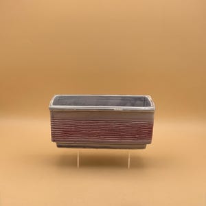 May include: Rectangular ceramic planter with a gray rim and a textured, reddish-brown band. The planter is elevated on clear supports against a solid tan background. Ideal for small plants or succulents.