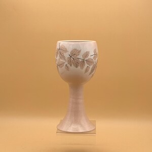 May include: A light pink ceramic goblet with a wide bowl and a slender stem. The bowl is decorated with a pattern of brown leaves. The goblet is displayed on a clear acrylic stand against a light orange background.