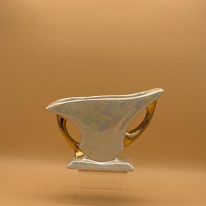 May include: A vintage, iridescent white ceramic vase with gold-colored handles. The vase has a unique shape with a wide opening and a rectangular base. The gold handles curve gracefully from the body.