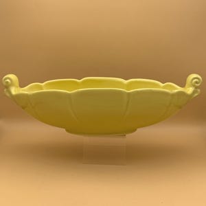 May include: A pale yellow ceramic bowl with a scalloped rim and two curled handles. The bowl has a wide, oval shape and sits on a clear stand. The bowl's color is a soft, muted yellow.