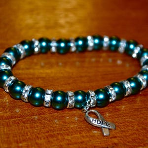 May include: Teal green beaded bracelet with silver accents and a silver charm in the shape of a ribbon with the word "HOPE" on it.