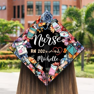 May include: A graduation cap with a black background, adorned with colorful floral patterns and medical-themed illustrations. The word "Nurse" is written in white script, along with "RN 202_" and a name. A tassel hangs from the cap.