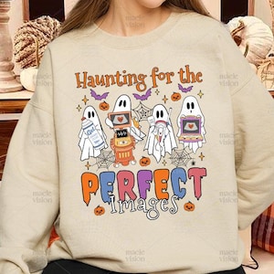 May include: Beige sweatshirt with the text "Haunting for the PERFECT Images" in orange, purple, and blue. The design features cartoon ghosts holding various items, including a laptop and a fire hydrant. Halloween-themed.