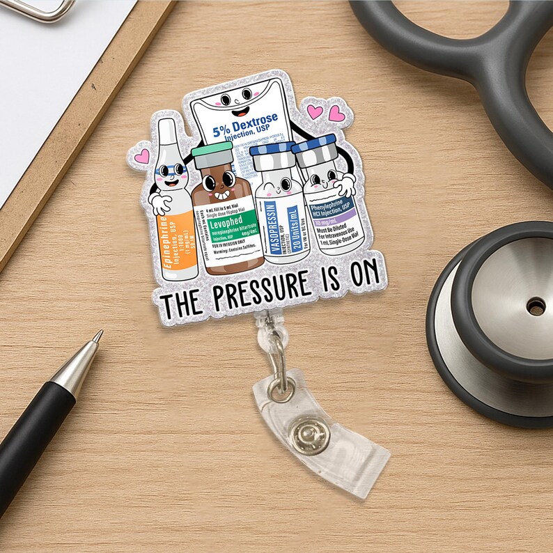 May include: A clear badge reel with a cartoon design featuring medicine bottles and the text "THE PRESSURE IS ON." The badge reel has a clear plastic strap and a metal clip. A stethoscope and pen are in the background.