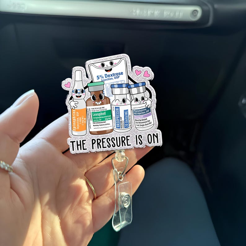 May include: A badge reel featuring cartoon illustrations of medicine vials and a bag of dextrose. The text "THE PRESSURE IS ON" is displayed below the illustrations. The reel has a clear plastic clip and a retractable cord.