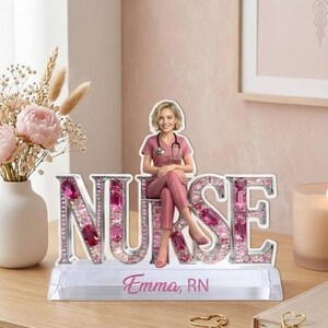 May include: A decorative nurse figurine with the word "NURSE" spelled out in silver letters, embellished with pink gems. A figurine of a nurse in pink scrubs sits atop the letters. The base reads "Emma, RN".