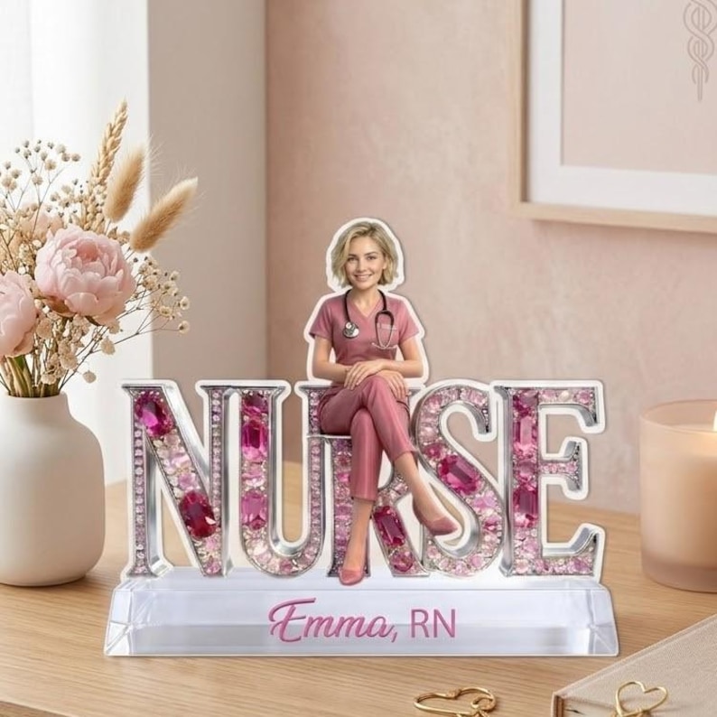 May include: A decorative nurse figurine with the word "NURSE" spelled out in silver letters, embellished with pink gems. A figurine of a nurse in pink scrubs sits atop the letters. The base reads "Emma, RN".