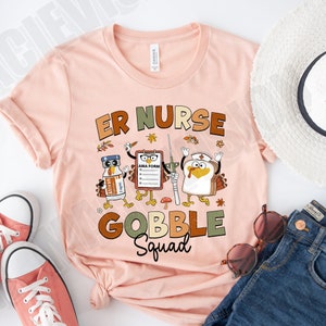 May include: A pink t-shirt with the text "ER Nurse Gobble Squad" in brown and green lettering. The design features a turkey, a bottle of saline solution, and a clipboard with an AMA form.