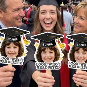 May include: A group of people at a graduation ceremony holding photo props. The props feature a child's face with a graduation cap, the name "Sophia", and the text "LOOK WHO'S GRADUATED". The graduate is wearing a black cap and gown.