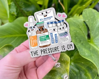 May include: A badge reel featuring cartoon illustrations of medical vials and a bag of dextrose. The text "THE PRESSURE IS ON" is displayed below the illustrations. The badge reel is silver and has a clear plastic clip.