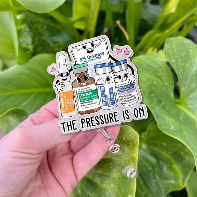 May include: A badge reel featuring cartoon illustrations of medical vials and a bag of dextrose. The text "THE PRESSURE IS ON" is displayed below the illustrations. The badge reel is silver and has a clear plastic clip.