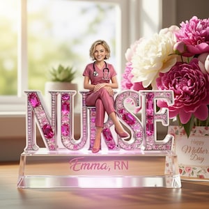 May include: A pink and silver Nurse figurine with the name "Emma, RN" on the base. The word "NURSE" is spelled out in large, jeweled letters. A woman in a pink nurse's uniform sits atop the letters. A bouquet of pink and white flowers is in the background.