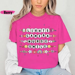 May include: A pink t-shirt with a graphic design that says "Lights Camera Nurse Smile" with colorful icons and flowers surrounding the text.