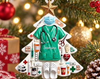 May include: A white Christmas tree-shaped ornament featuring a green scrub top with the name Jasmine, a stethoscope, a blue face mask, a gold star, and various medical items including syringes, pills, a heart, an ambulance, and white clogs.