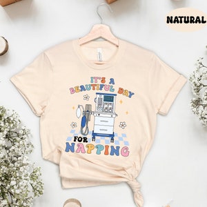 May include: A light beige t-shirt with a graphic design of a medical ventilator with the text "It's a beautiful day for napping" in rainbow colors.