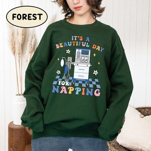 May include: Green sweatshirt with a colorful graphic design that says "It's a beautiful day for napping". The graphic features a medical ventilator and flowers.