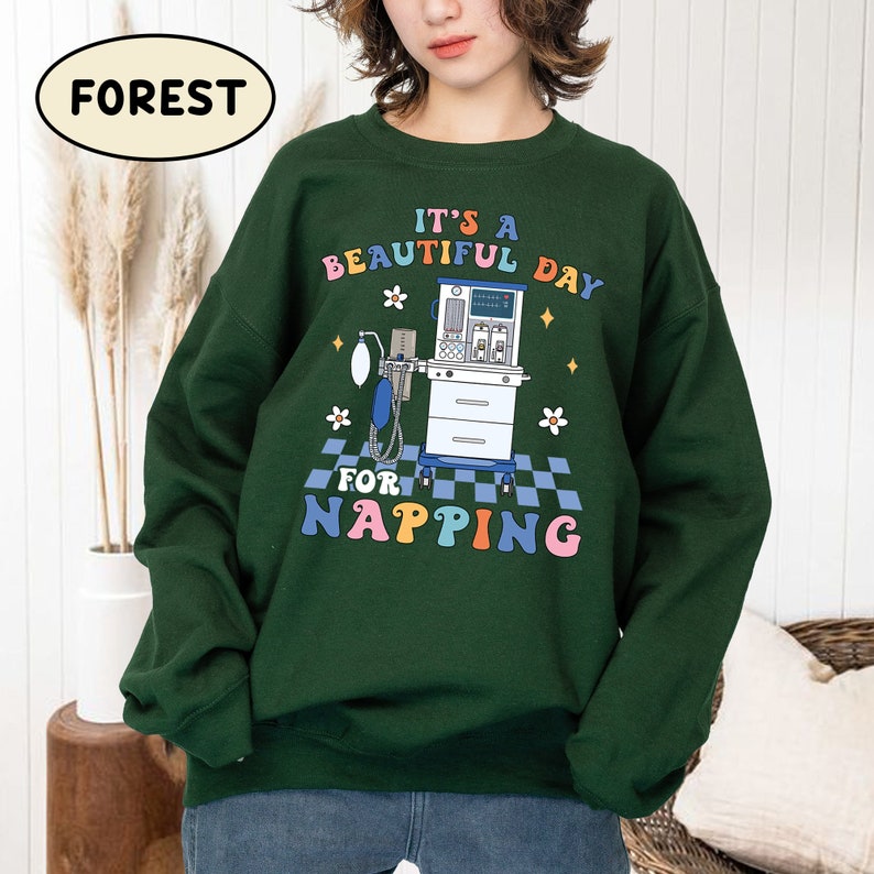 May include: Green sweatshirt with a colorful graphic design that says "It's a beautiful day for napping". The graphic features a medical ventilator and flowers.